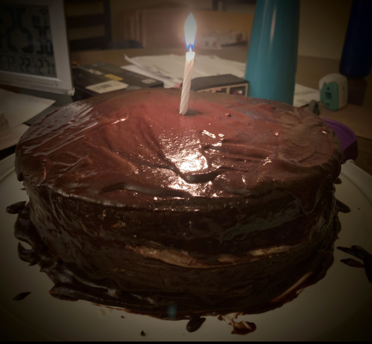 Homemade chocolate layer cake with one lit candle in the center. A messy dining room table is partially obscured by a shadow effect.